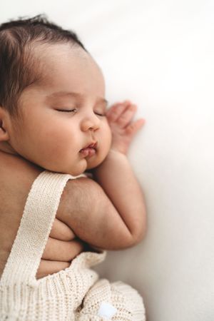 Peaceful sleeping newborn baby in cream knit overalls lying on a white fluffy surface.