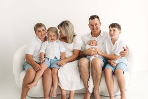 Happy family with newborn baby and three boys in white outfits sitting on a white couch.