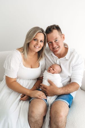 Smiling couple in white outfits holding their swaddled newborn baby on a white couch.