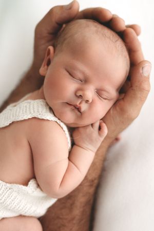 Sleeping newborn baby in white knit outfit cradled gently in father's hands.