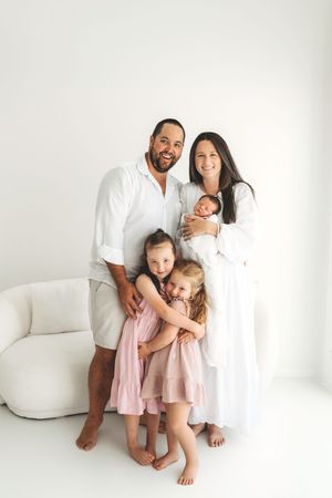 Happy family of five with parents in white, three daughters in pink, posing in bright white studio.