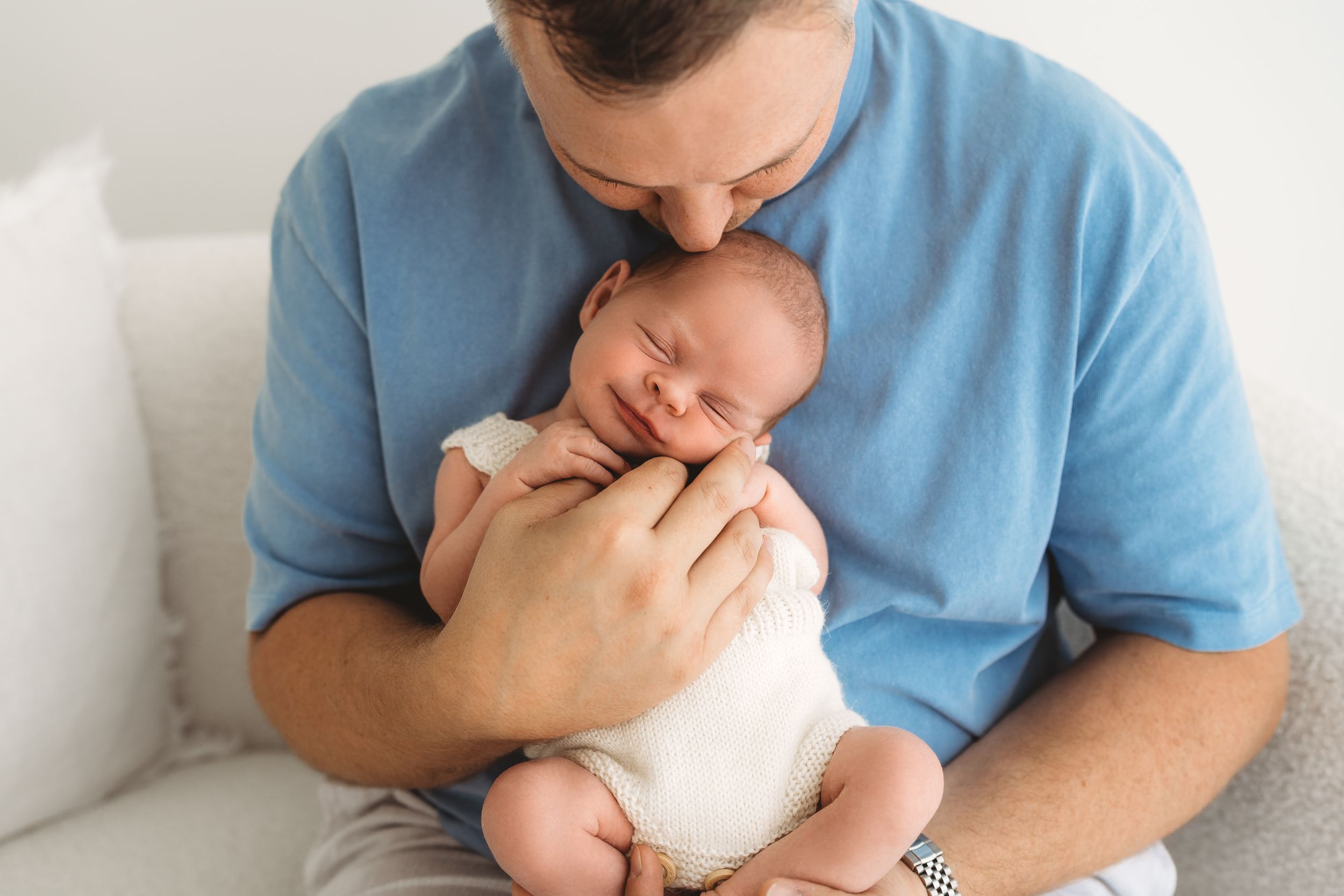 Father in blue shirt kissing sleeping newborn baby held against his chest.
