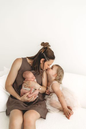 Mother in brown dress holding newborn while toddler daughter in pink tutu gives a kiss.