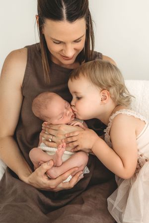 Mother smiling as blonde toddler daughter kisses sleeping newborn baby on white background.