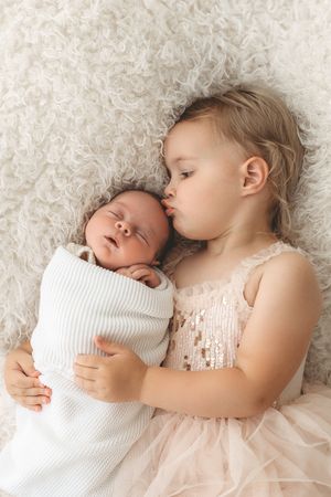 Toddler girl in pink dress kissing sleeping newborn sibling wrapped in white on fluffy cream blanket.