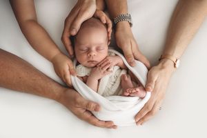 Parents hands cradling sleeping newborn baby wrapped in white cloth on white background.