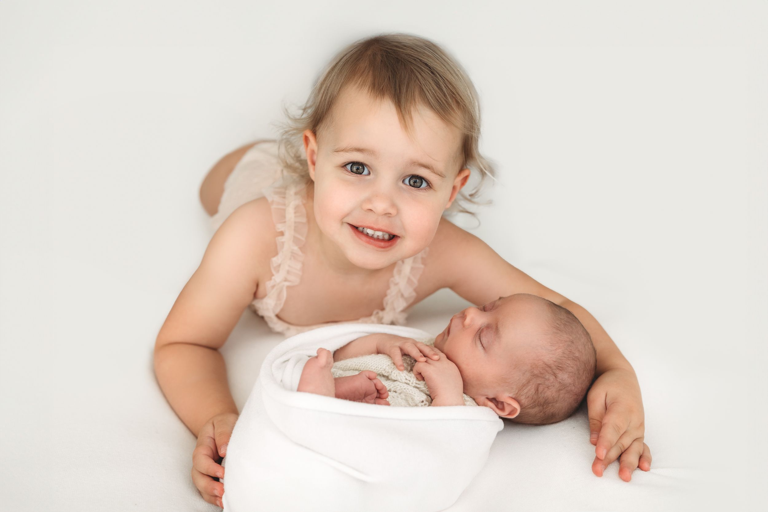 Smiling toddler girl in pink dress posing with sleeping newborn sibling wrapped in white.