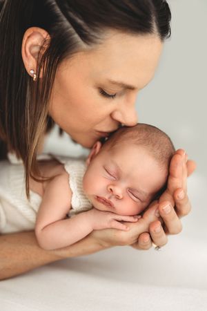 Mother tenderly kissing sleeping newborn baby held gently in her hands on white background.