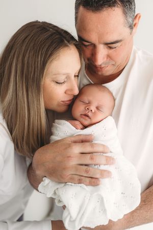 Loving parents kissing and holding sleeping swaddled newborn baby in white outfits.
