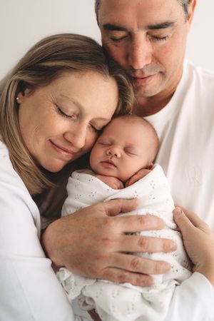 Happy couple smiling and cuddling sleeping swaddled newborn baby in white clothing.