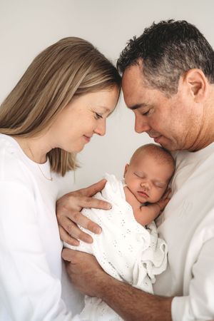 Smiling parents touching foreheads together while holding sleeping swaddled newborn baby.