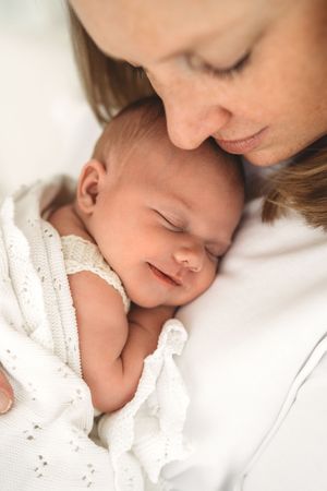 Tender close-up of mother nuzzling smiling sleeping newborn baby wrapped in white knit.