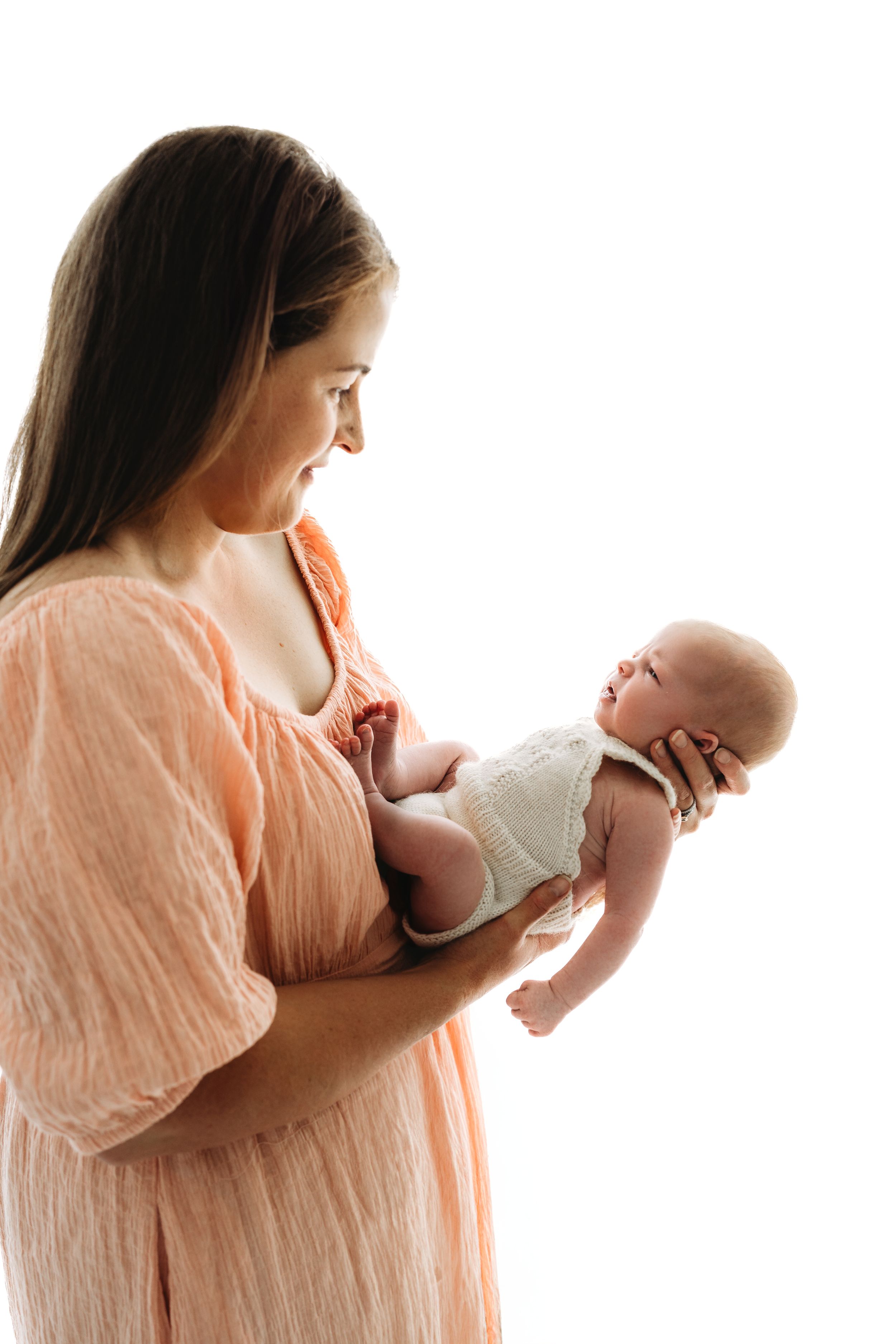 Smiling mother in peach dress holding and gazing at newborn baby on white background.