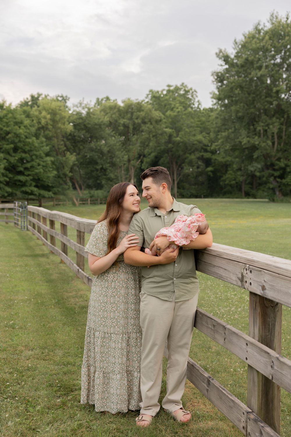 A couple stands with their newborn baby against a wooden fence in a serene outdoor setting with green trees.
