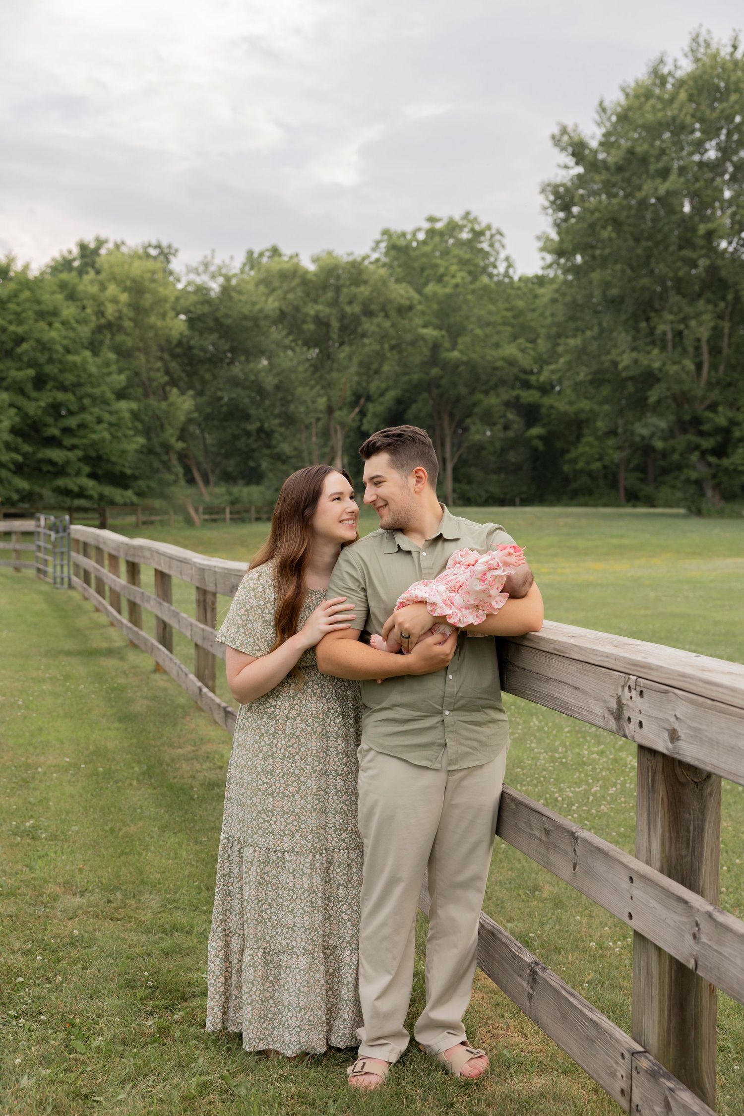 A couple stands with their newborn baby against a wooden fence in a serene outdoor setting with green trees.