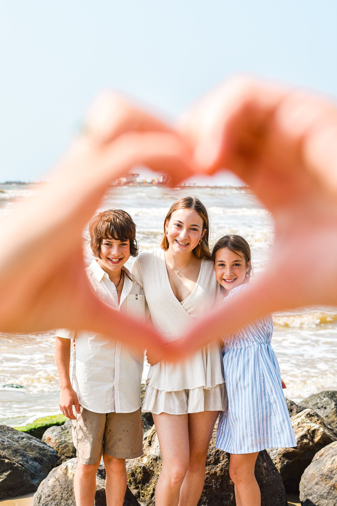 Heart-shaped hands frame three people posing together on a rocky beach with ocean waves in the background.