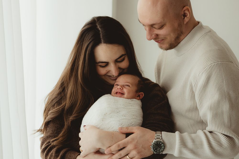 Parents lovingly embrace their newborn baby swaddled in white blanket by window with soft natural light.