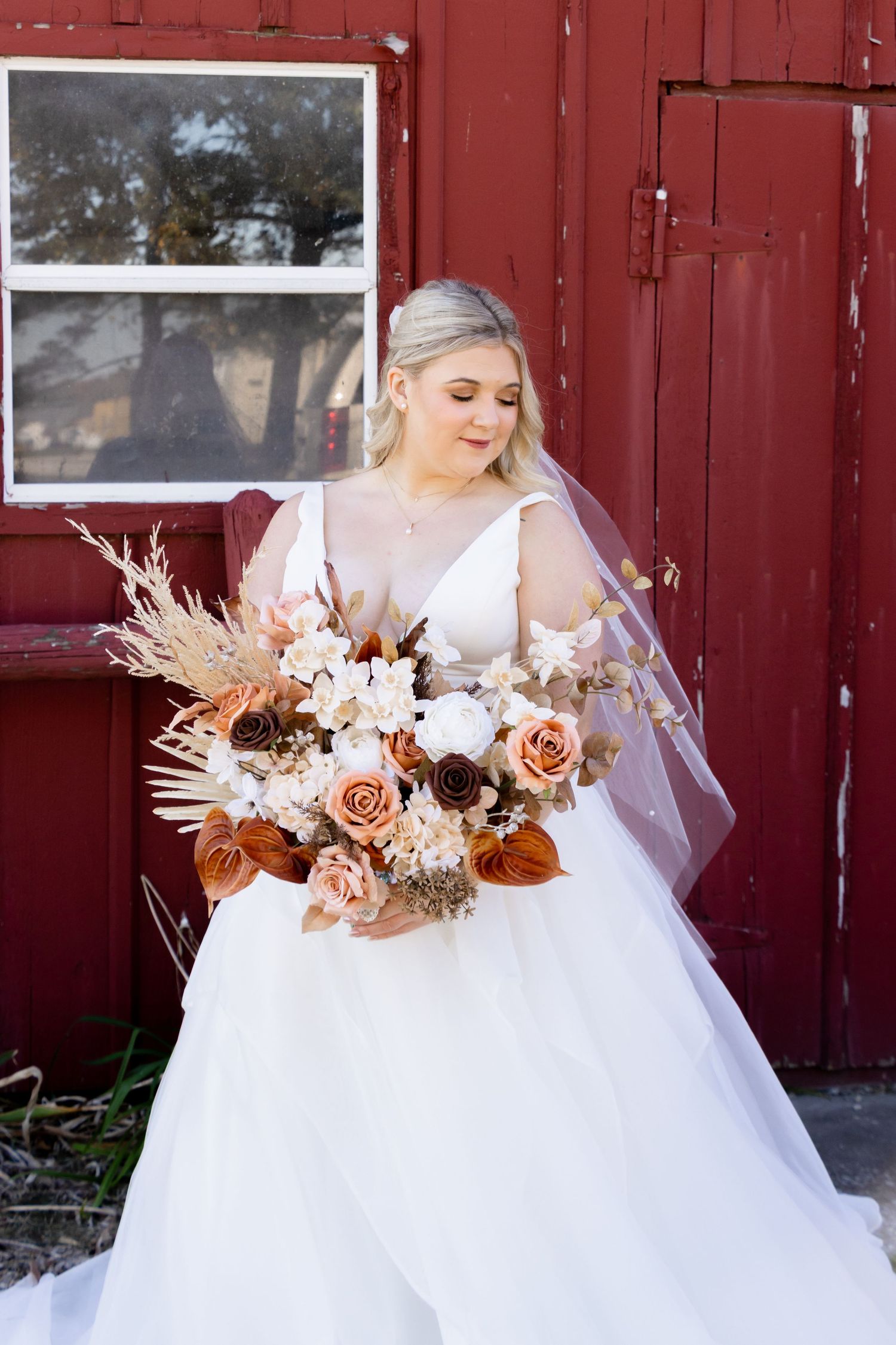Bride in white gown with dried flower bouquet against red barn backdrop.