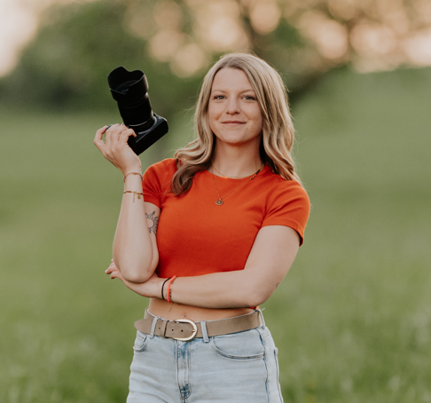 Frau in rotem T-Shirt und heller Jeans posiert selbstbewusst im Freien vor einem natürlichen Bokeh-Hintergrund.