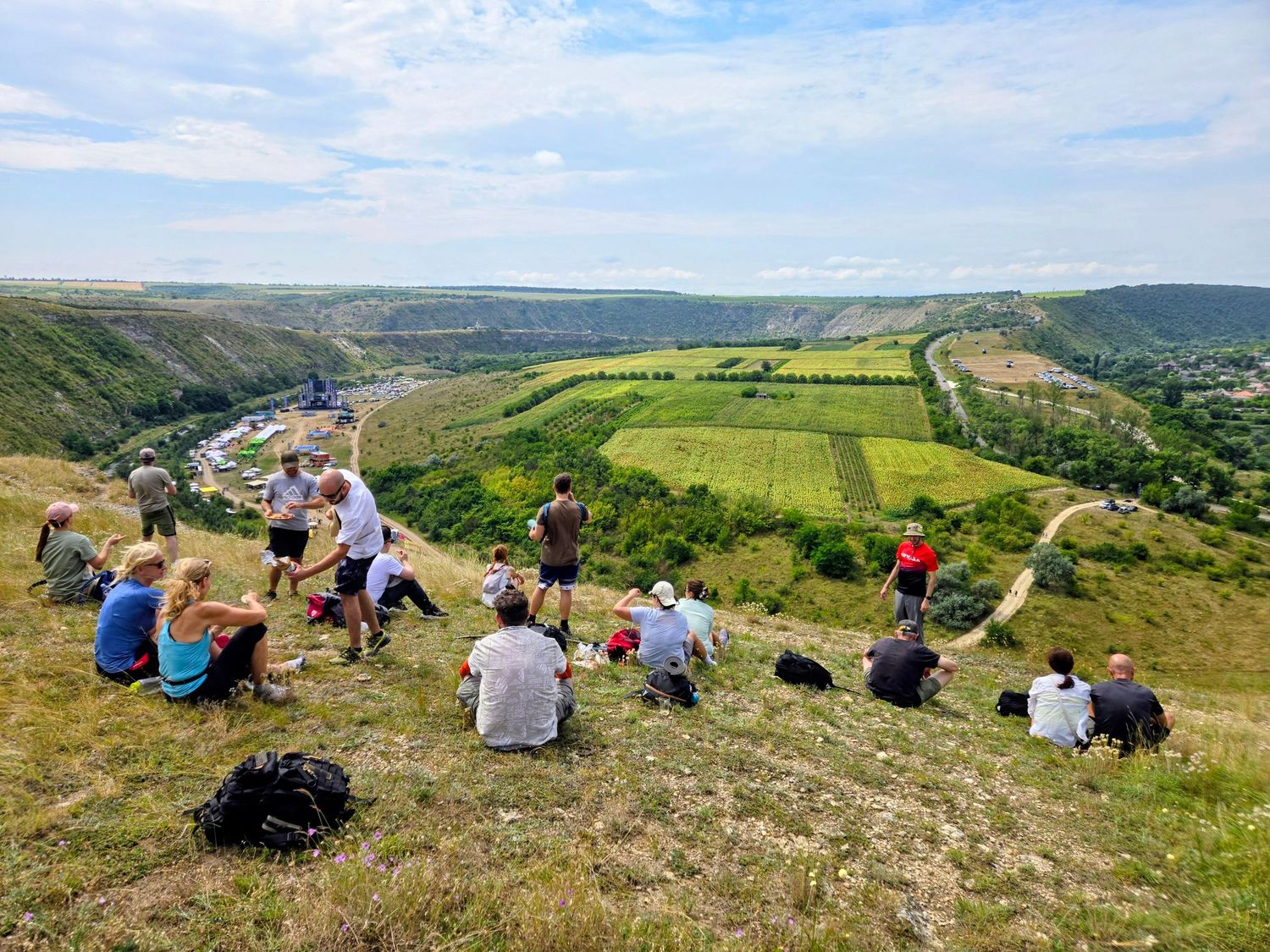 Group of hikers resting on a scenic hilltop overlooking a lush green valley with winding paths below.
