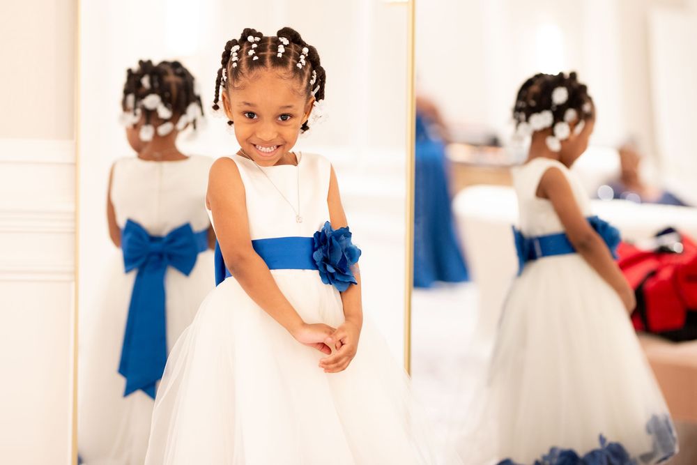 Young flower girl in white dress with blue sash smiling at mirror reflection.