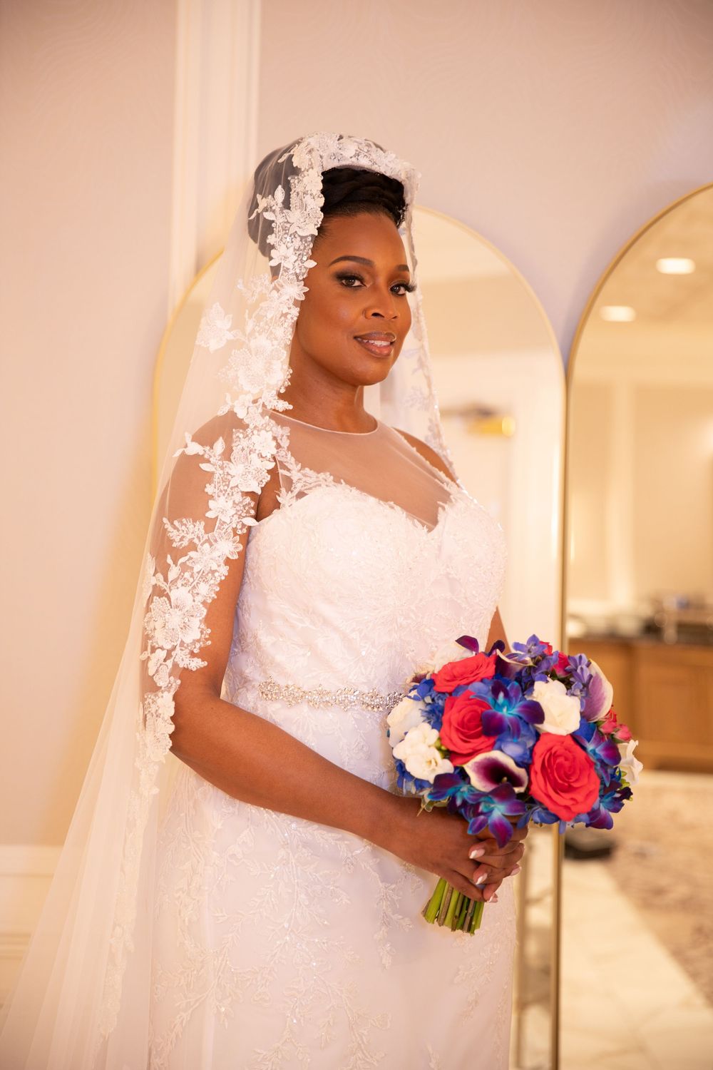 A bride holds a vibrant red and blue bouquet while wearing a white lace dress and veil.