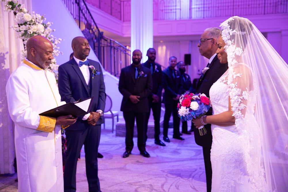 Wedding ceremony in a purple-lit ballroom with white columns and chandeliers.