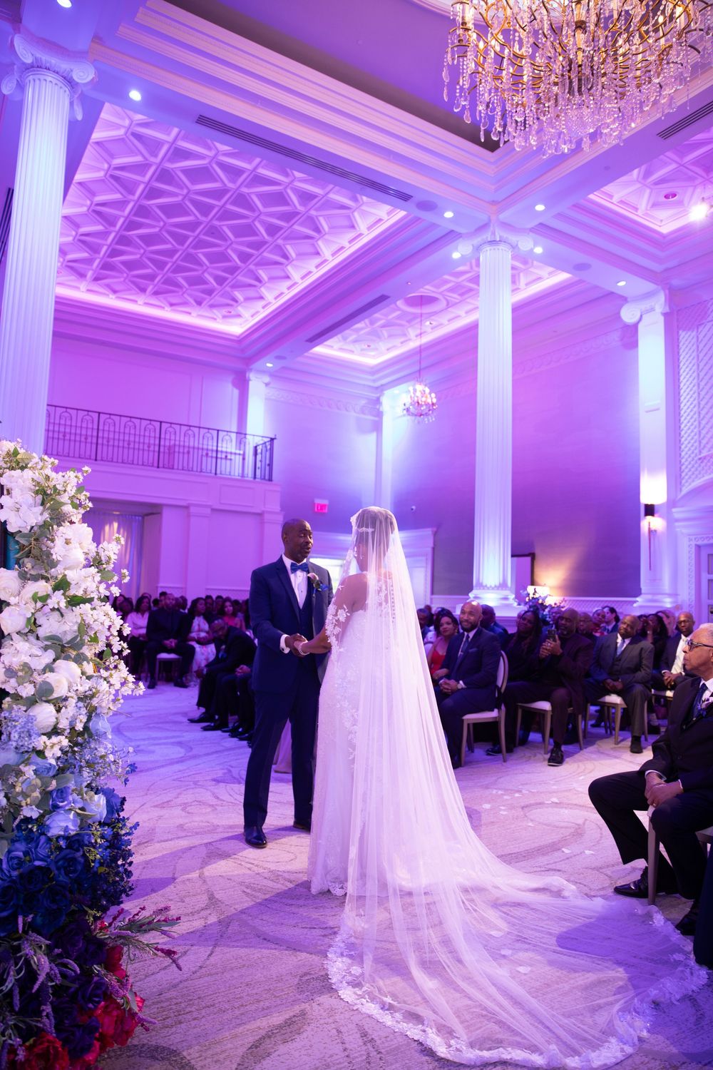 Elegant ballroom wedding venue with purple uplighting, chandeliers, and white columns.