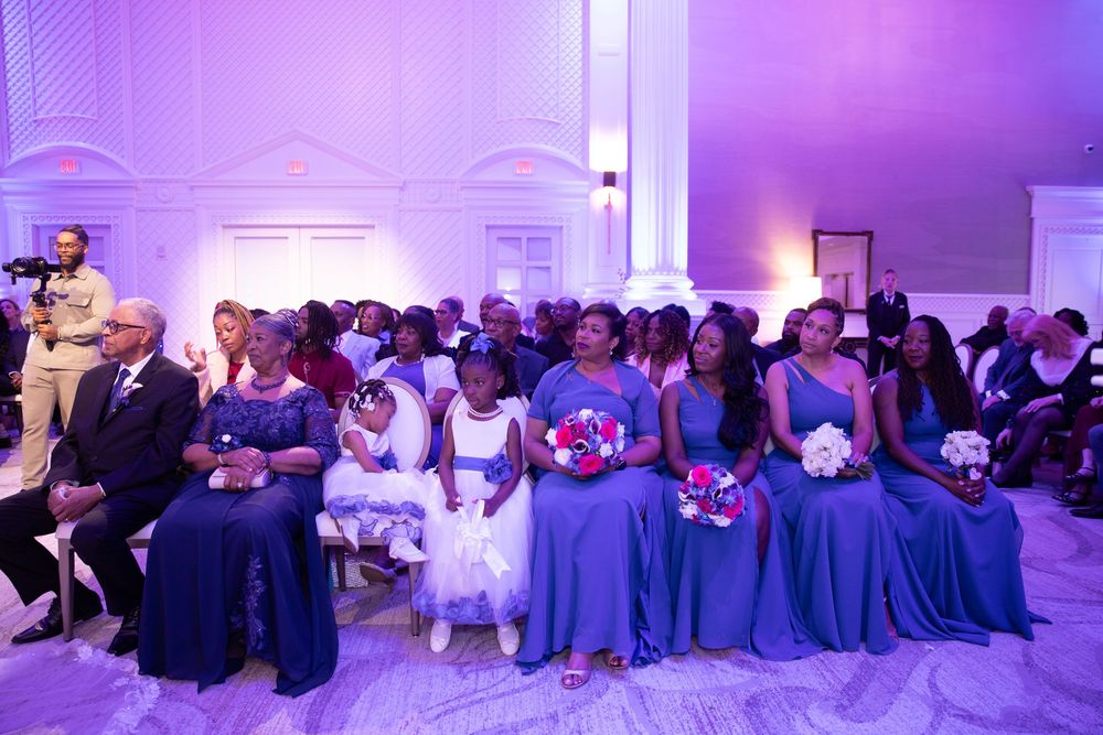 Wedding guests seated in rows during ceremony under purple lighting.