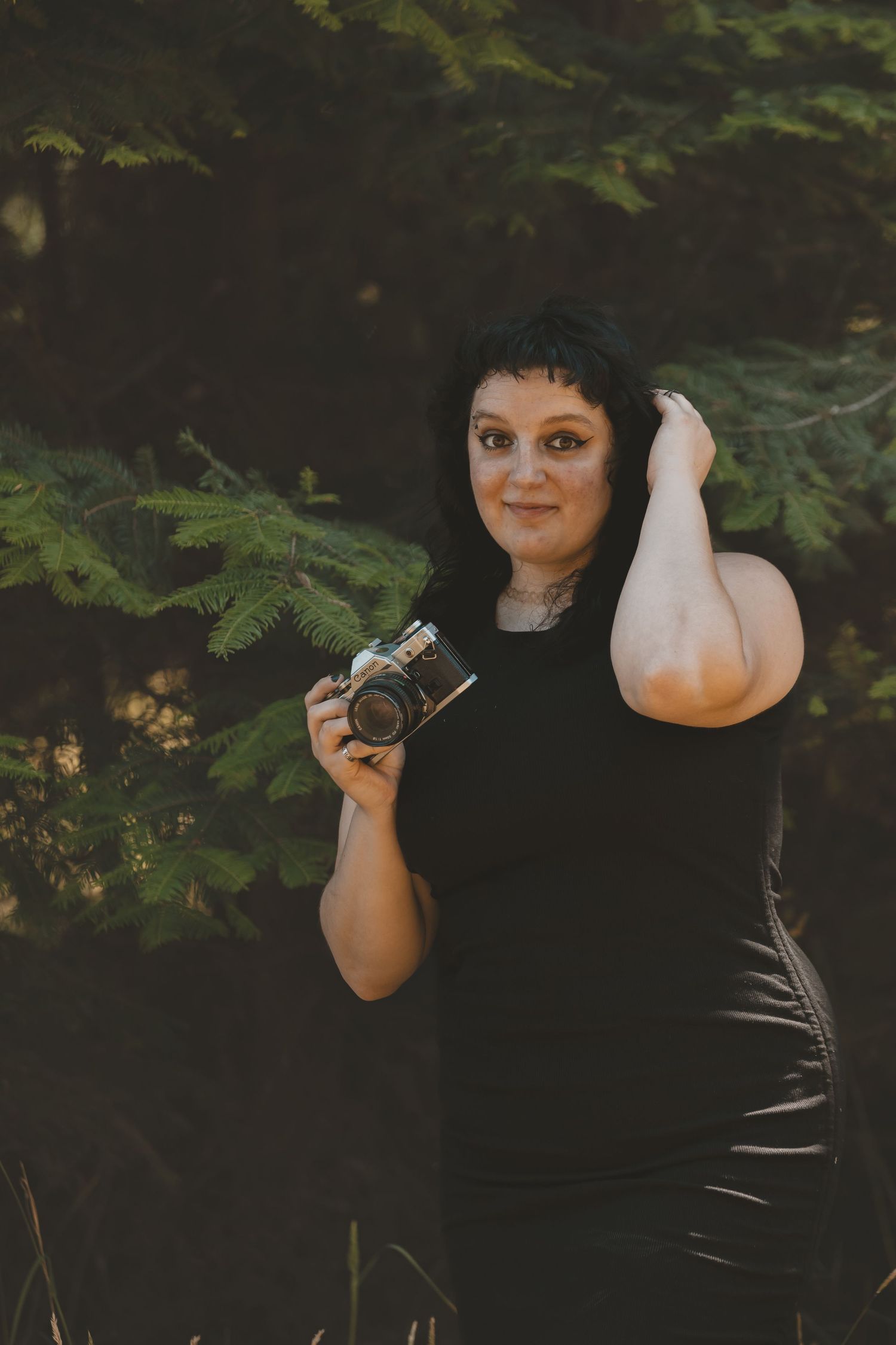 A woman standing in a field of daisys with a tree behind her. She is holding a camera. She is wearing checkered pants and a black shirt.