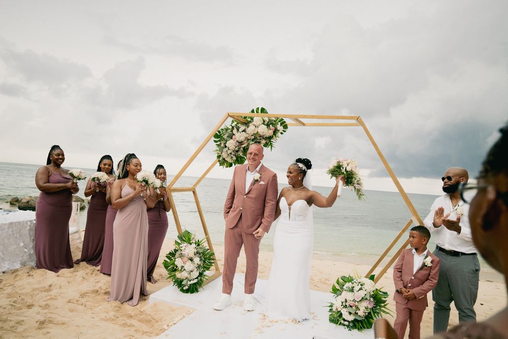 A wedding ceremony takes place on a beach under a hexagonal arch decorated with white and pink flowers.