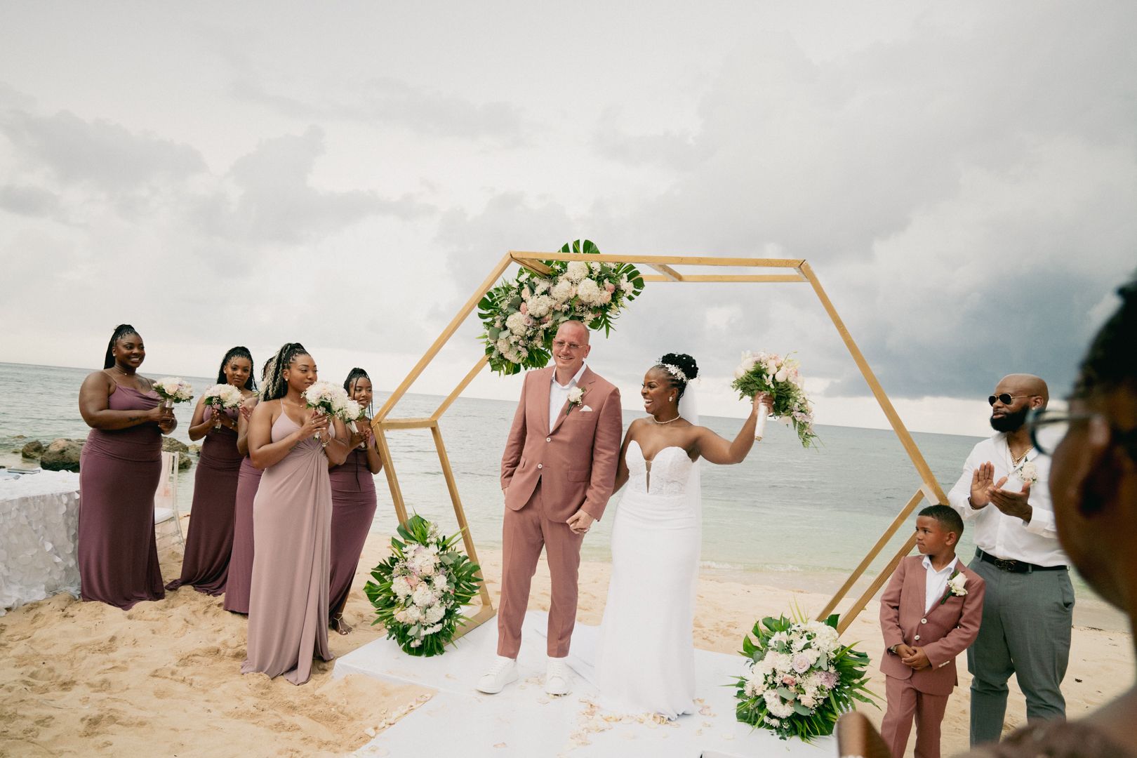 A wedding ceremony takes place on a beach under a hexagonal arch decorated with white and pink flowers.