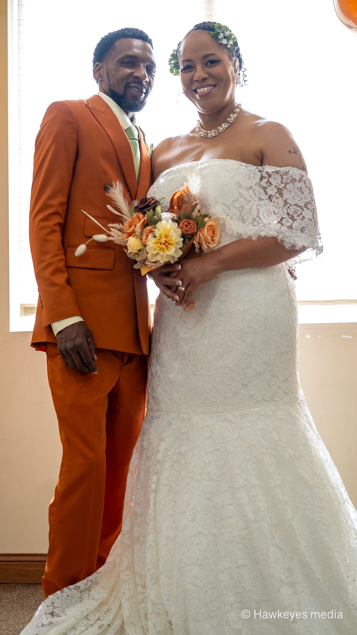 A couple poses on their wedding day, with the groom in a rust-colored suit and bride in a white lace gown holding orange flowers.