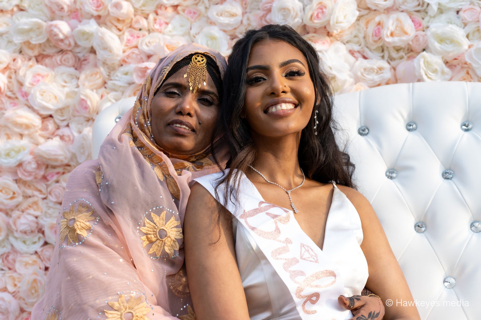 Two women in white outfits pose together against a pink floral backdrop with a white tufted chair.