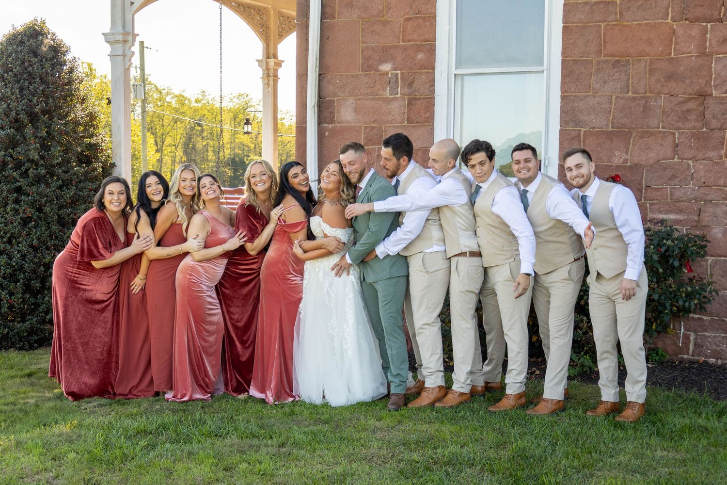 Wedding party in burgundy and tan attire posed outside brick church building.
