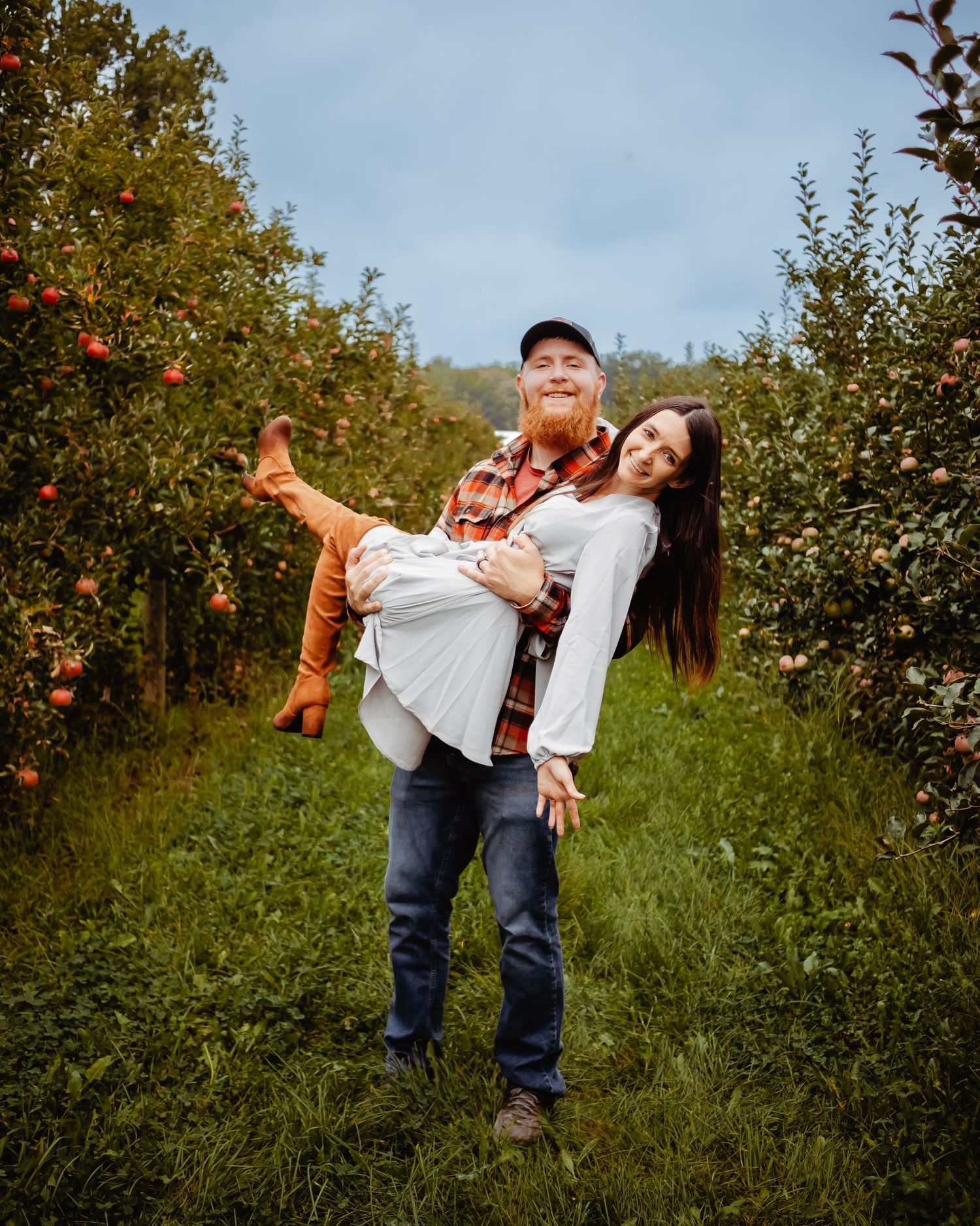A couple shares a playful moment in an apple orchard during autumn with colorful trees in the background.