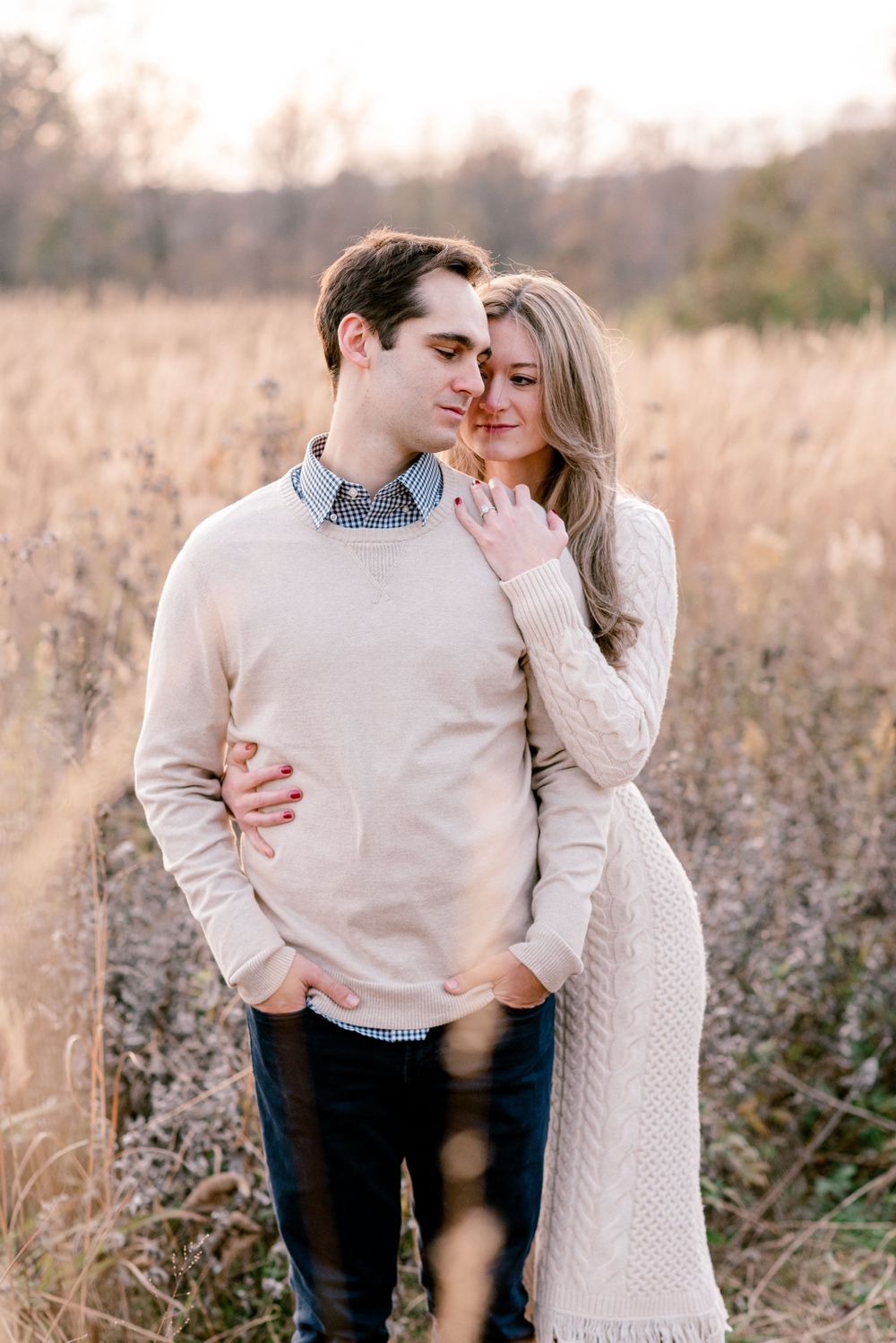 A couple embraces in a sunlit autumn field wearing neutral colored sweaters.
