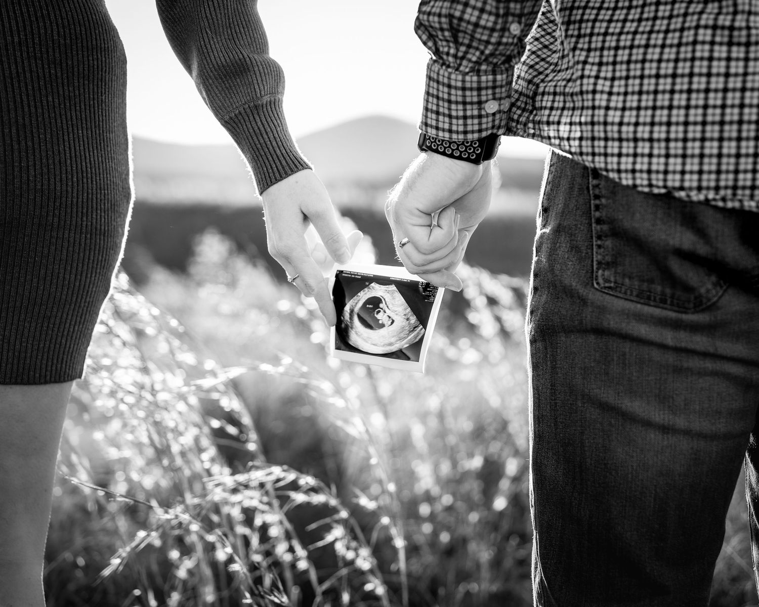 A couple holds a sonogram photograph in a sunlit field of wildflowers, captured in black and white.