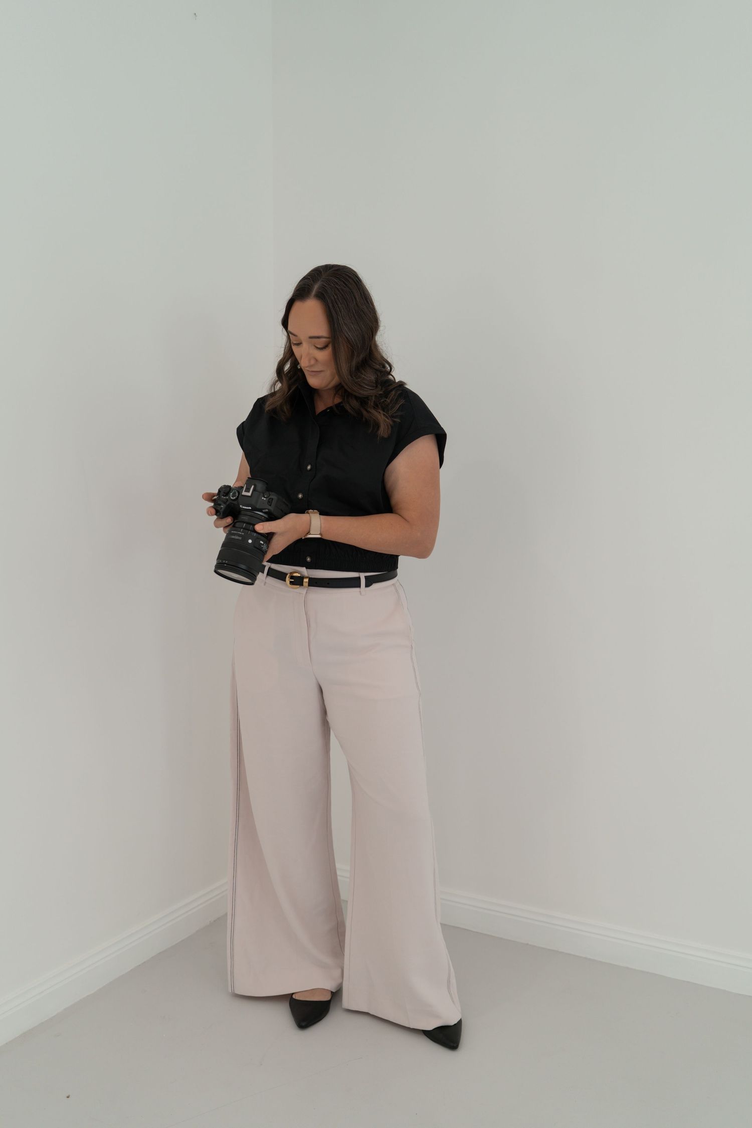 A photographer dressed in a black top and cream wide-leg pants poses in different angles against a white studio backdrop, admiring her camer