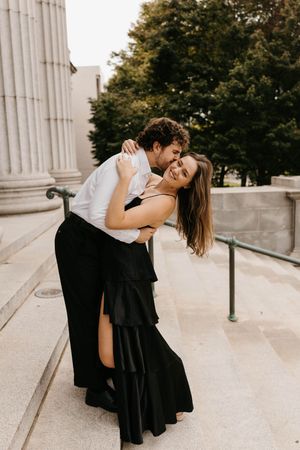 Man dipping woman in black dress while kissing her cheek on outdoor stone steps.
