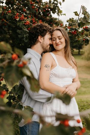 Couple embracing tenderly under red berry branches during outdoor engagement photography session.