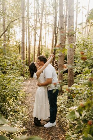 Couple sharing a romantic kiss on a sunlit forest trail during engagement session.