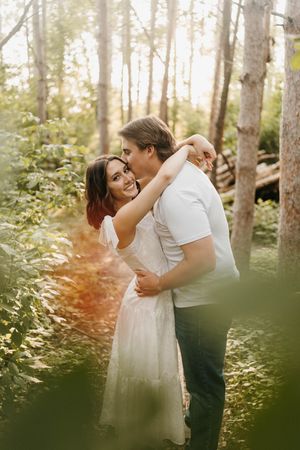 Couple embracing and smiling in a sunlit forest, woman in white dress with arms around man in white shirt.