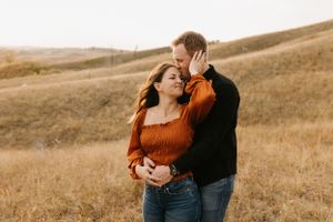 Couple in golden prairie field, woman in rust top leaning into man in black sweater at sunset.