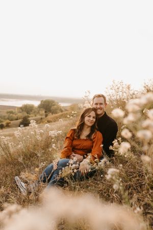 Couple sitting together in wildflower meadow with scenic lake view, woman in rust blouse smiling warmly.