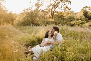 Couple sitting in golden meadow grass at sunset, woman in white dress leaning back against man.