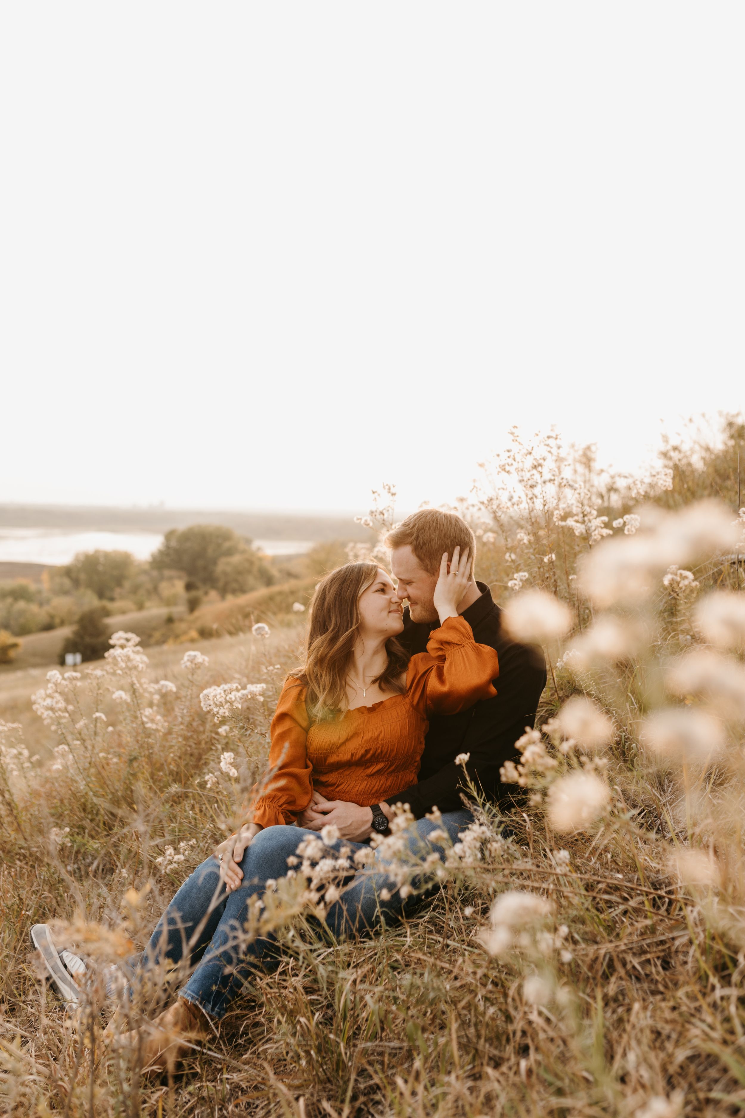 Couple sharing a kiss among wildflowers in a meadow, woman in orange top, scenic lake backdrop.