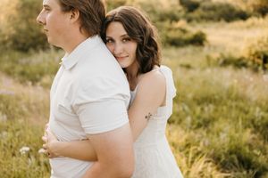 Woman in white dress hugging man from behind in a sunny green field, smiling at camera.