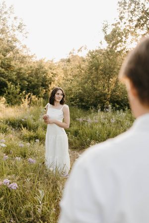 Woman in white dress standing in wildflower field at golden hour, facing man in foreground.