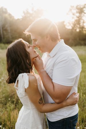 Couple about to kiss in sunlit meadow, backlit by golden sunset light, embracing closely.
